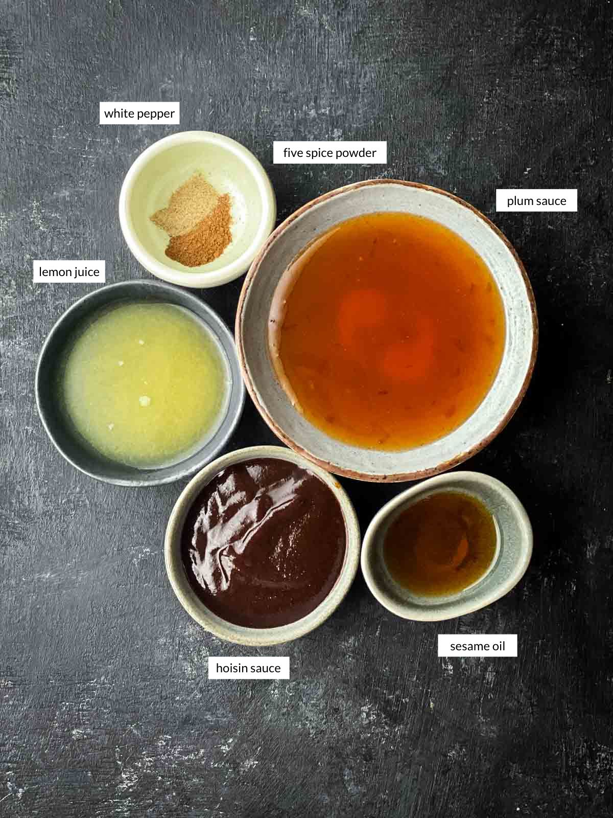 Overhead view of small bowls containing plum sauce, hoisin sauce, lemon juice, sesame oil, Chinese five spice powder, and white pepper — ingredients for Yee Sang plum sauce salad dressing.