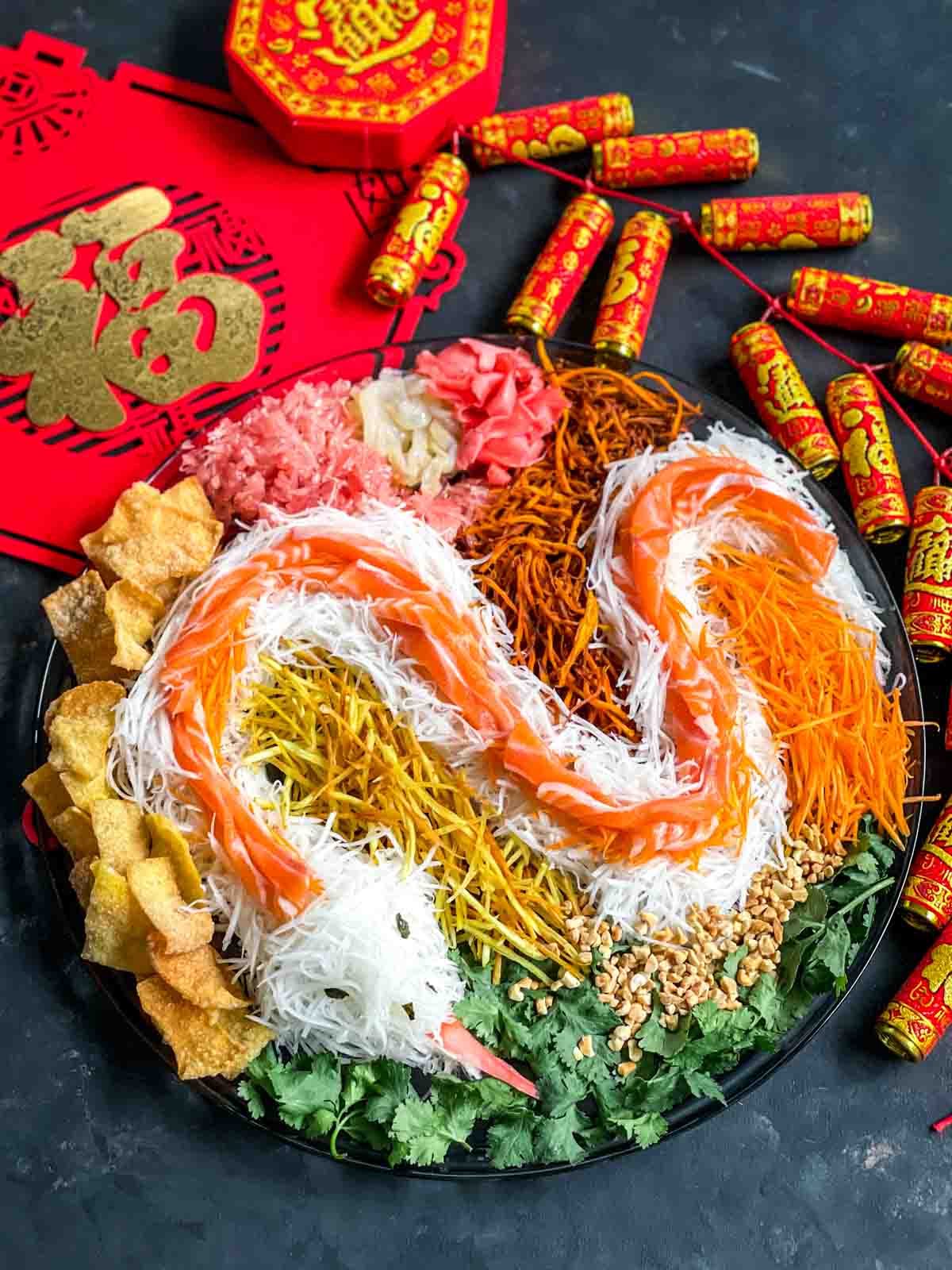 Chinese New Year Yee Sang salad on a large plate with the shredded vegetables made in the shape of a snake for the year of the snake. Surrounded by Chinese red packets and firecracker decorations.