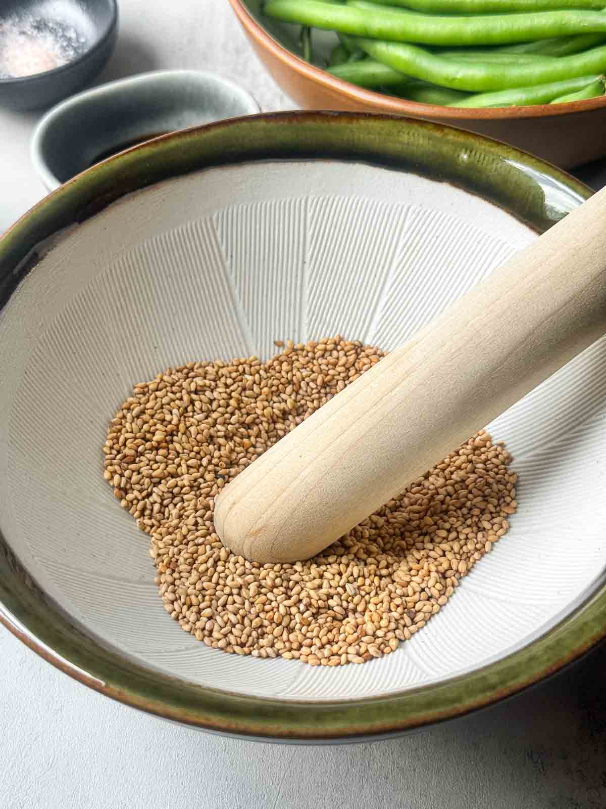 Grinding toasted sesame seeds in a traditional Japanese suribachi mortar with a wooden pestle to prepare the base for gomae sesame sauce.