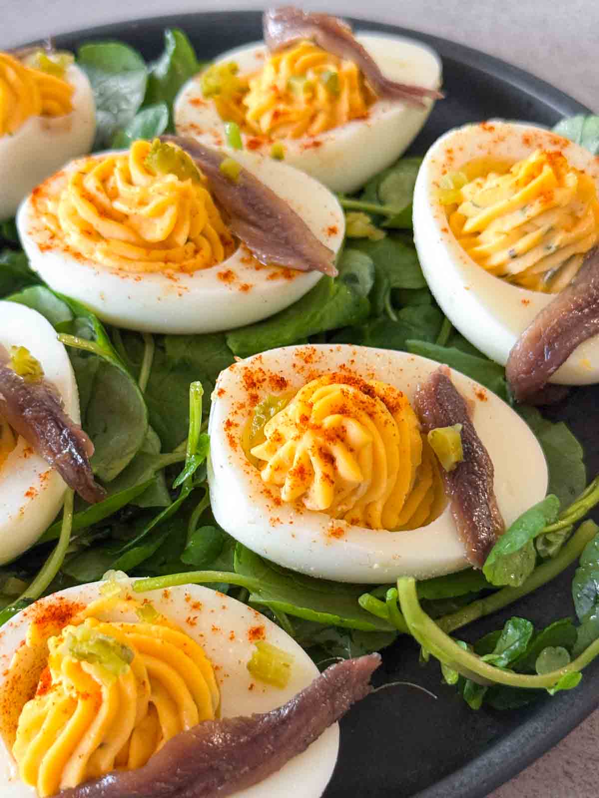 Overhead view of deviled eggs topped with anchovies on a bed of fresh watercress. The eggs are stuffed with creamy yolk filling and dusted with paprika — a make-ahead deviled egg appetizer served on a black plate.