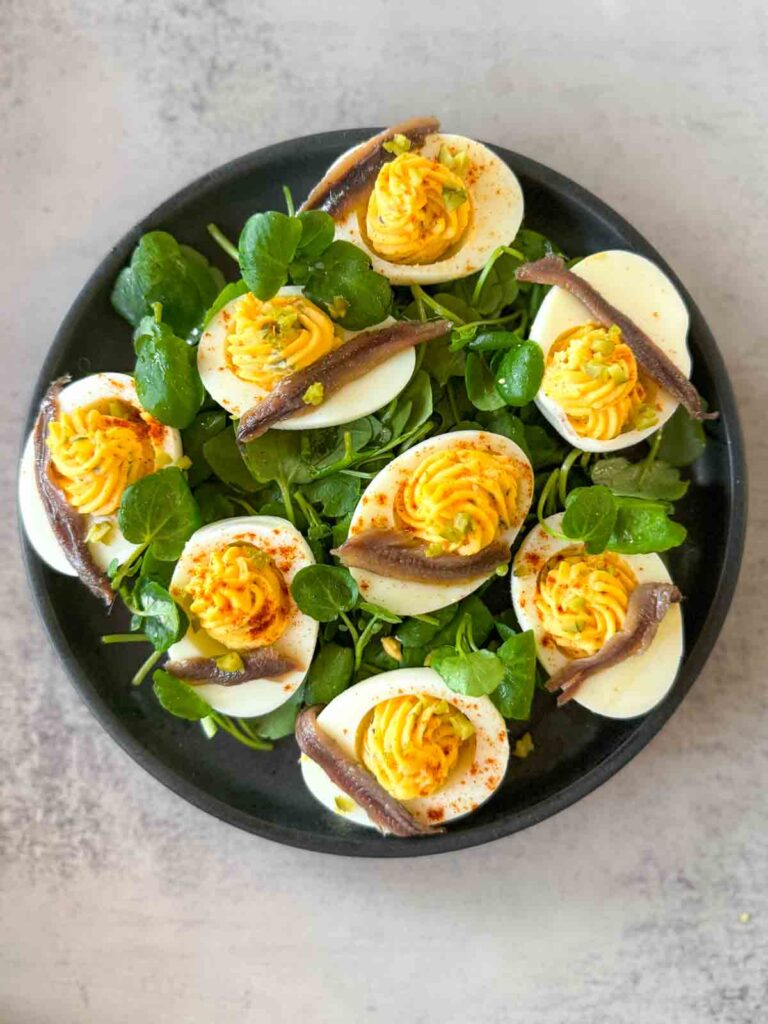 Overhead view of deviled eggs topped with anchovies on a bed of fresh watercress. The eggs are stuffed with creamy yolk filling and dusted with paprika — a make-ahead deviled egg appetizer served on a black plate.
