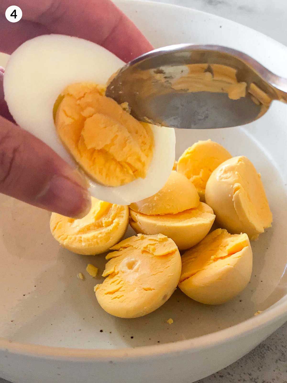 Scooping out firm egg yolks with a spoon from boiled egg halves in a white bowl, ready to mix for deviled egg stuffing.