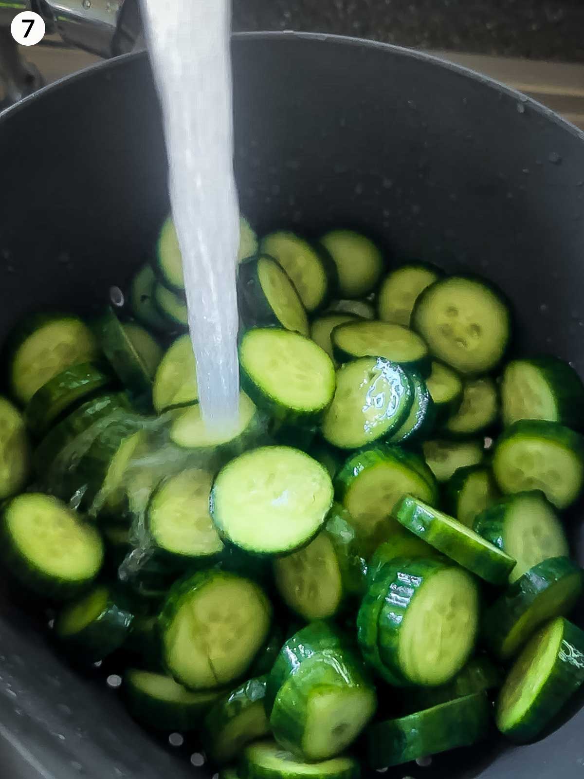 Cucumber slices being rinsed under cold water in a colander after salting — key step for crunchy cucumber kimchi.