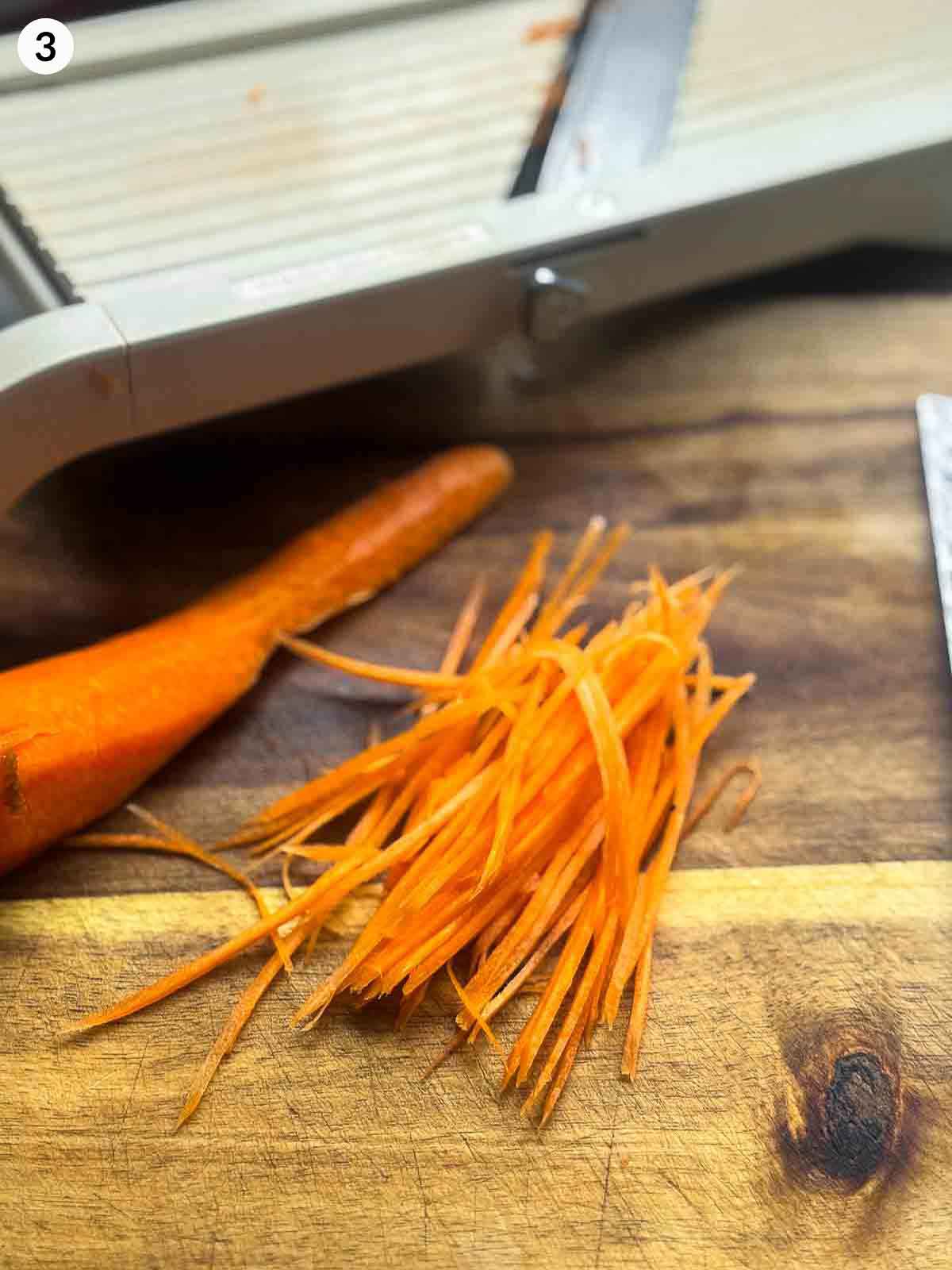 Thin julienned carrots on a wooden board beside a mandoline slicer — step-by-step preparation for Korean cucumber kimchi (Oi Kimchi).