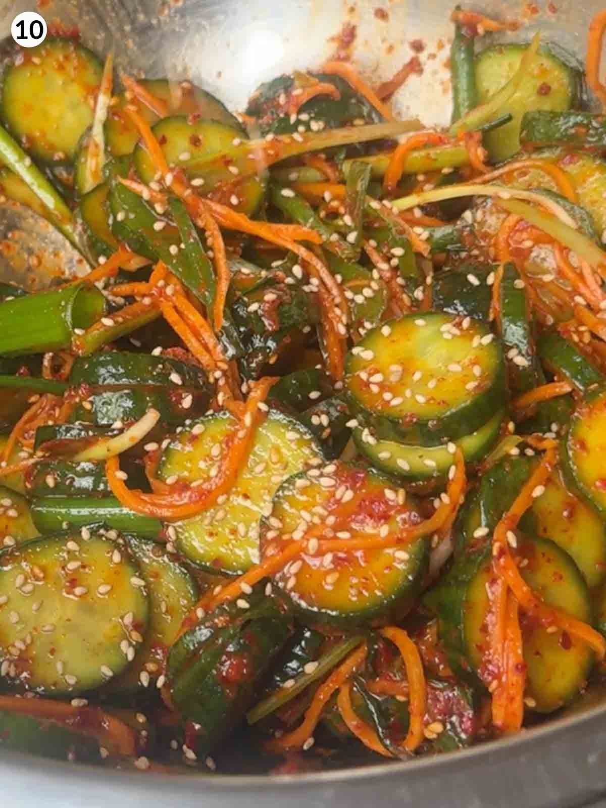 Sliced cucumbers, carrots, and scallions tossed with spicy gochugaru paste and sesame seeds in a metal bowl — the final step of making Korean cucumber kimchi (Oi Kimchi).