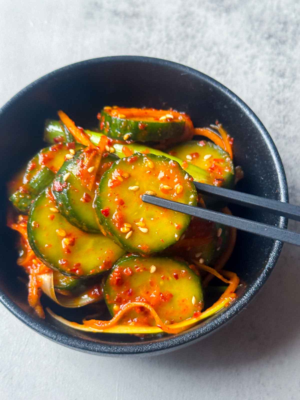 A small black bowl of Korean cucumber kimchi (Oi Kimchi) made with cucumber slices, shredded carrot, red chili flakes, and sesame seeds — a quick, spicy Korean side dish.
