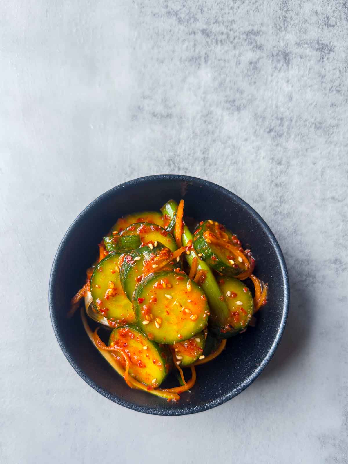 A small black bowl of Korean cucumber kimchi (Oi Kimchi) made with cucumber slices, shredded carrot, red chili flakes, and sesame seeds — a quick, spicy Korean side dish.