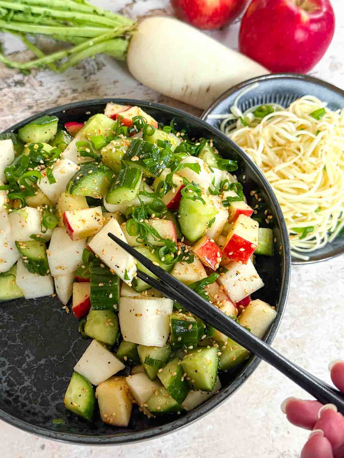 Daikon Cucumber Apple Salad with Togarashi Dressing on a black plate served with noodles. Daikon and apples in t he background