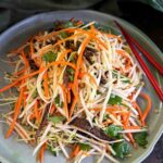 Close-up of Asian slaw with shredded kohlrabi, carrot, strips of beef jerky, fresh coriander leaves, snow pea sprouts, and crushed peanuts in a grey bowl.