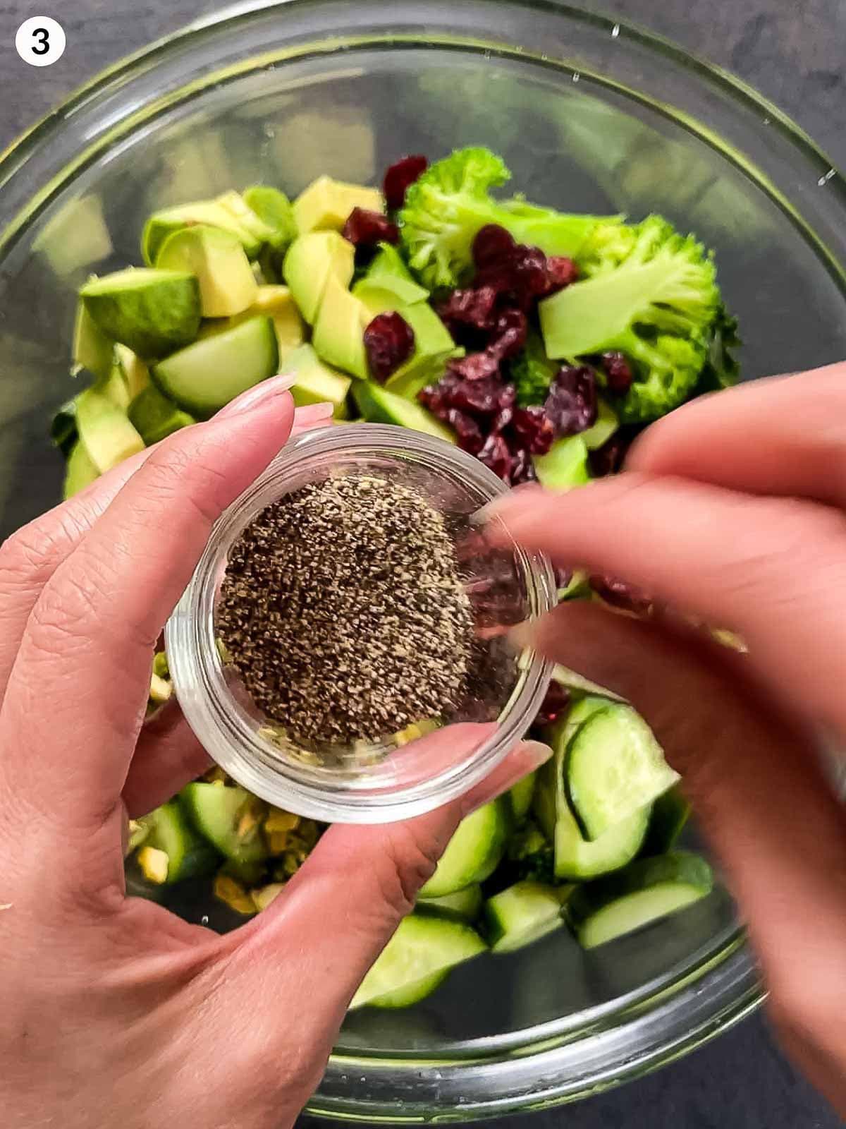 2 hands adding a pinch of pepper into a clear mixing bowl of Cranberry and Kale salad ingredients