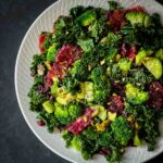 Cranberry and kale salad with blood orange wheels, blanched broccoli florets, diced avocado, dried cranberries, and chopped pistachios on a white ribbed plate against a dark background.