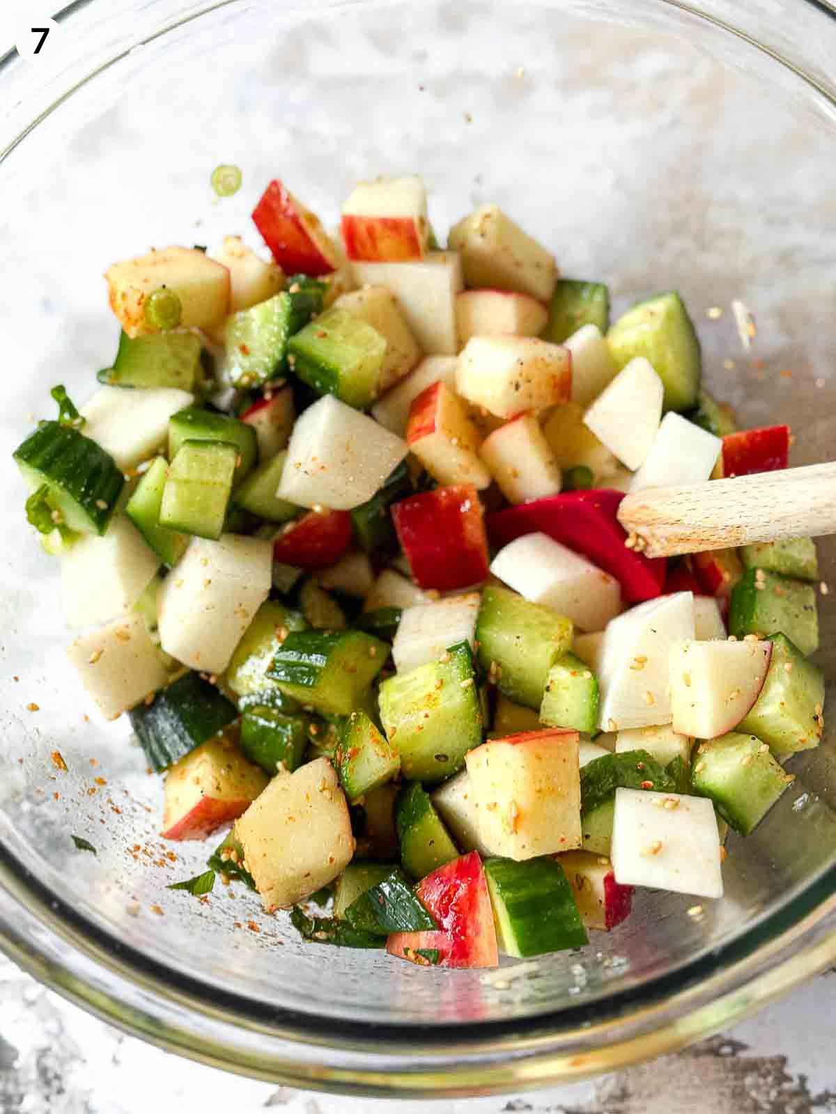 Tossing ingredients for Daikon Cucumber Apple Salad in a clear bowl with a red spatula