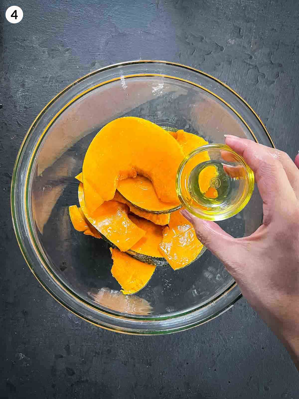 Adding olive oil to pumpkin wedges in a clear mixing bowl against a dark background