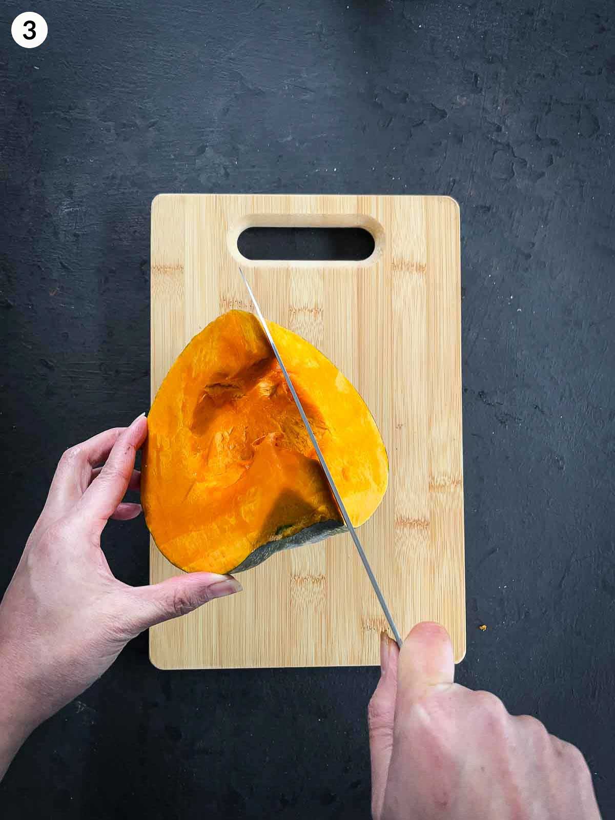 2 hands cutting kent pumpkin into wedges with a knife on a wooden chopping board against a dark background