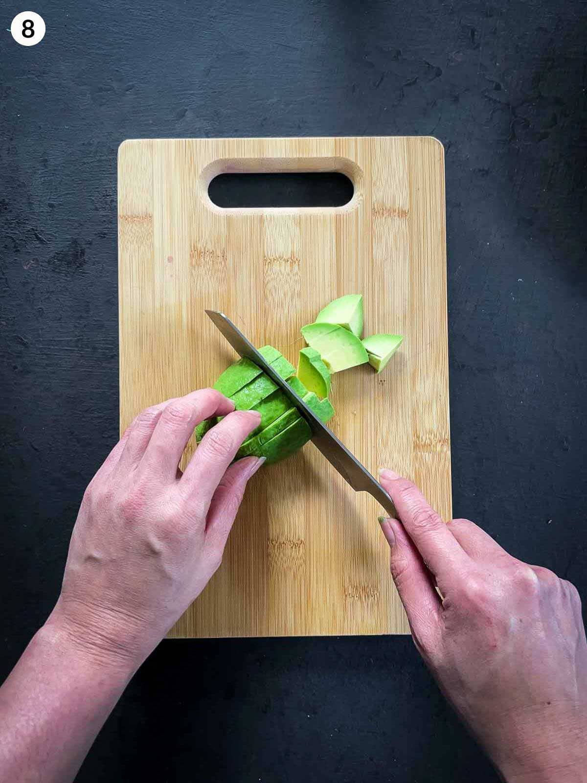 Hands dicing a peeled avocado on a wooden chopping board using a chef's knife, showing scored avocado being sliced into cubes against a dark background.