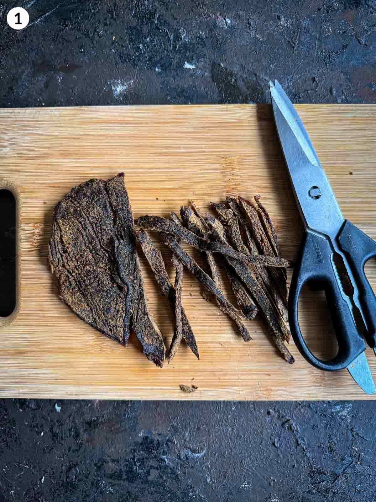 Using kitchen scissors to cut strips of beef jerky on a wooden board against a dark background