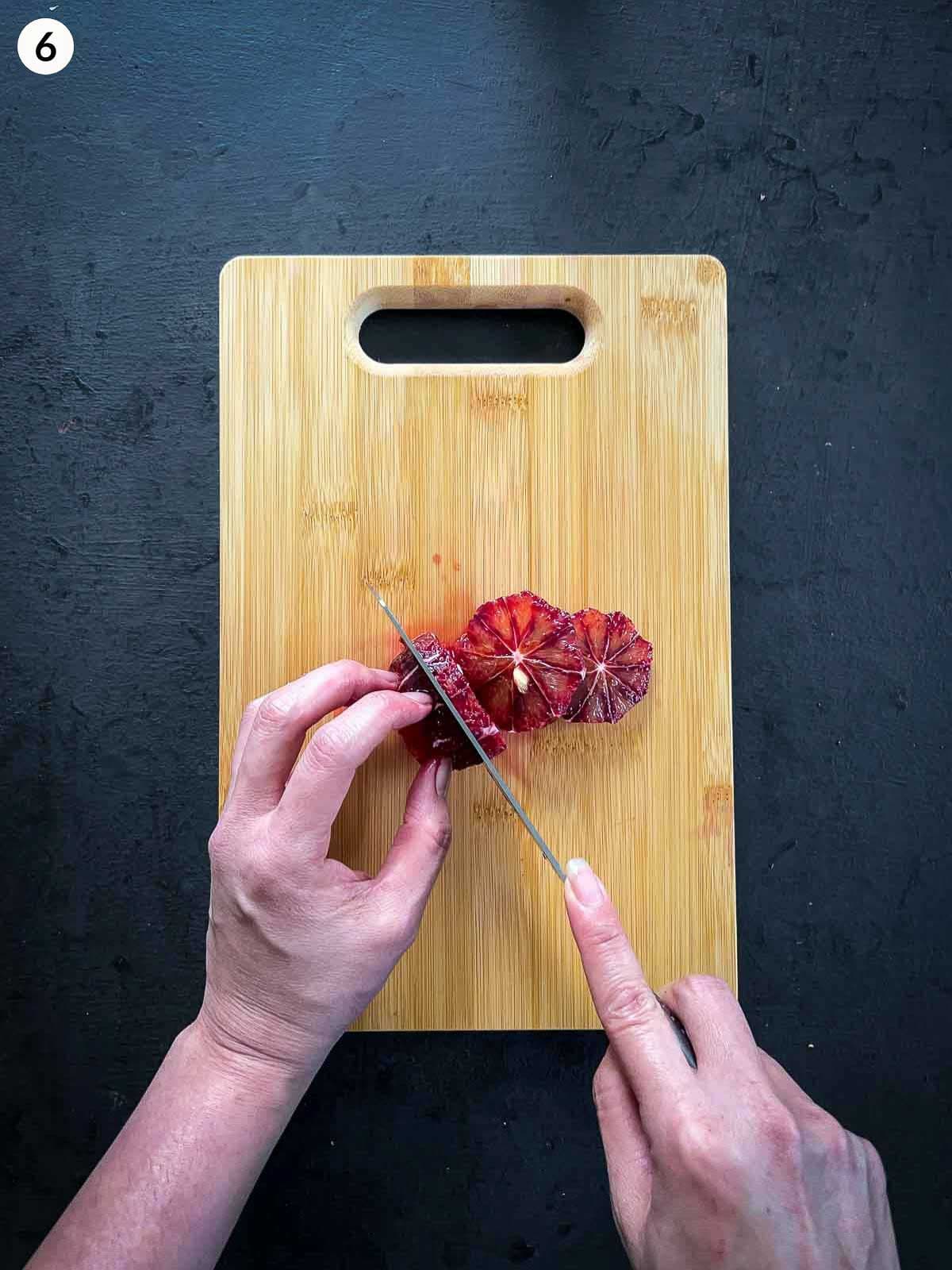 Hands slicing blood orange wheels on a wooden chopping board using a chef's knife, against a dark background.