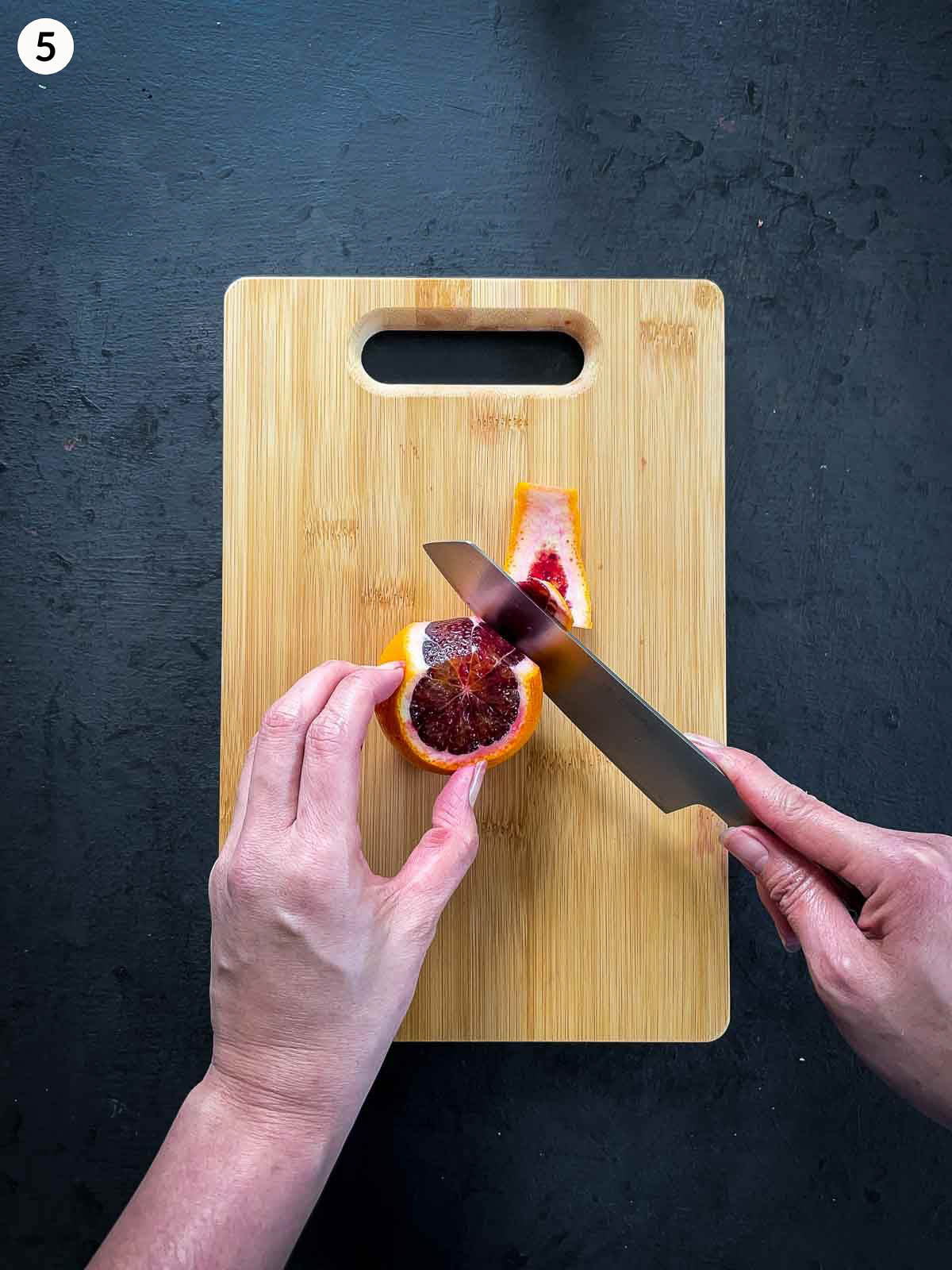 Hands slicing blood orange peel on a wooden chopping board using a chef's knife, against a dark background.