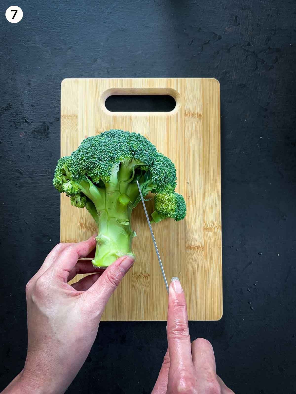 Hands cutting broccoli florets on a wooden chopping board using a chef's knife, against a dark background.
