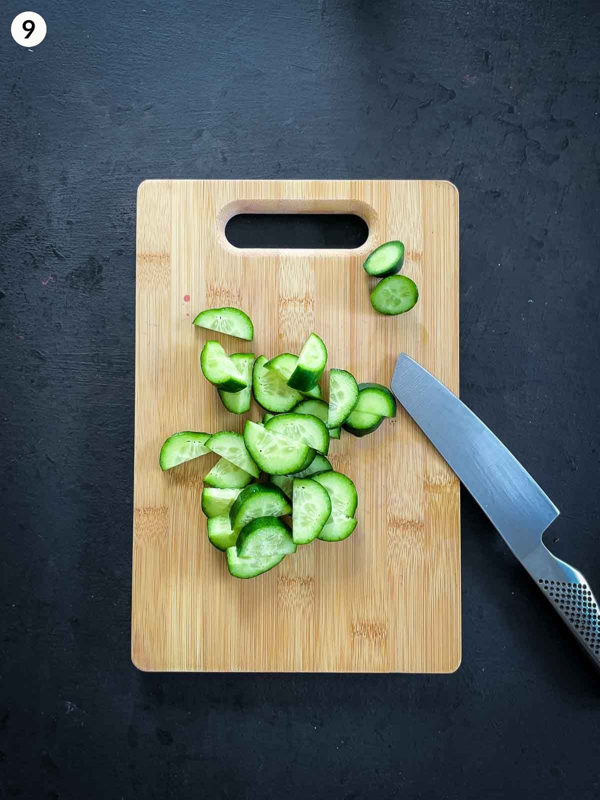Hands cutting cucumber on a wooden chopping board using a chef's knife, against a dark background.