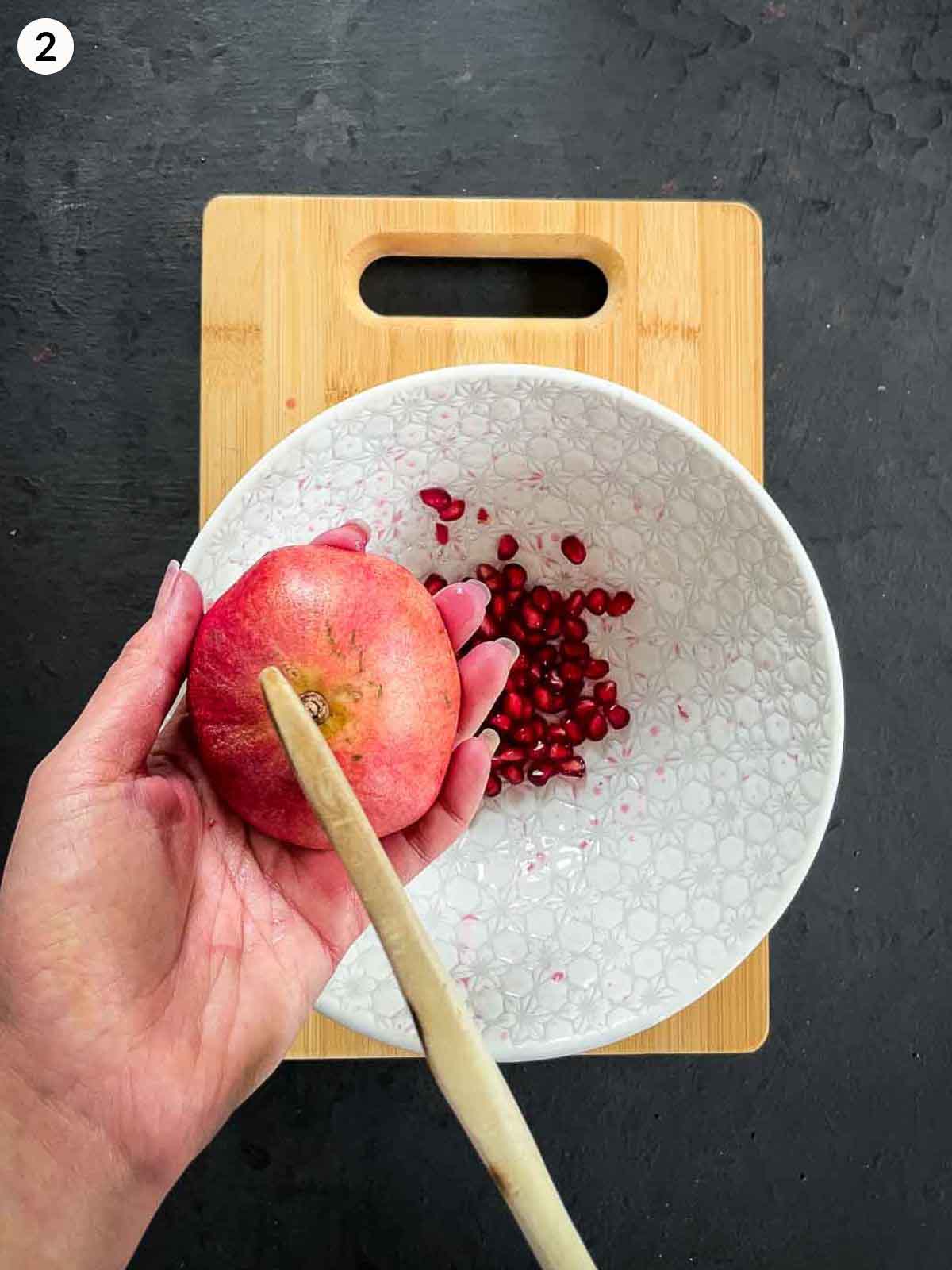 deseeding pomegranate with a wooden spoon into a small white bowl on a wooden board