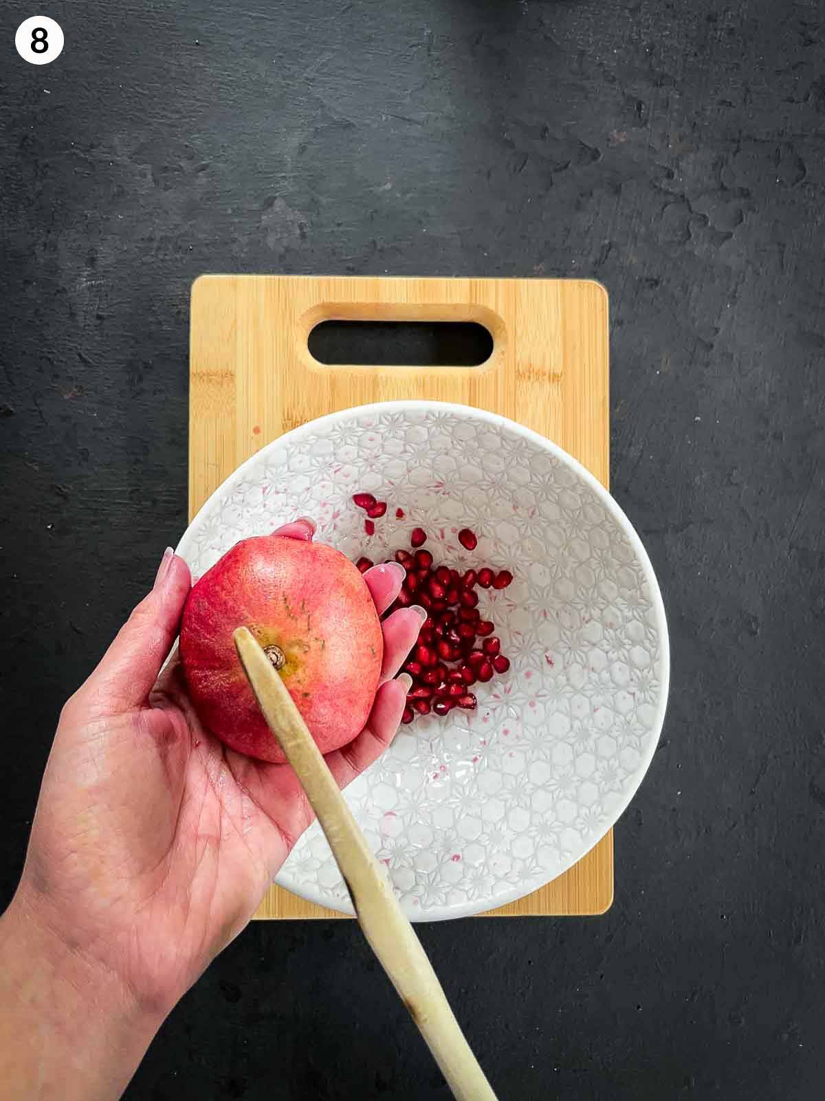 2 hands deseeding pomegranate with a wooden spoon on a wooden chopping board against a dark background