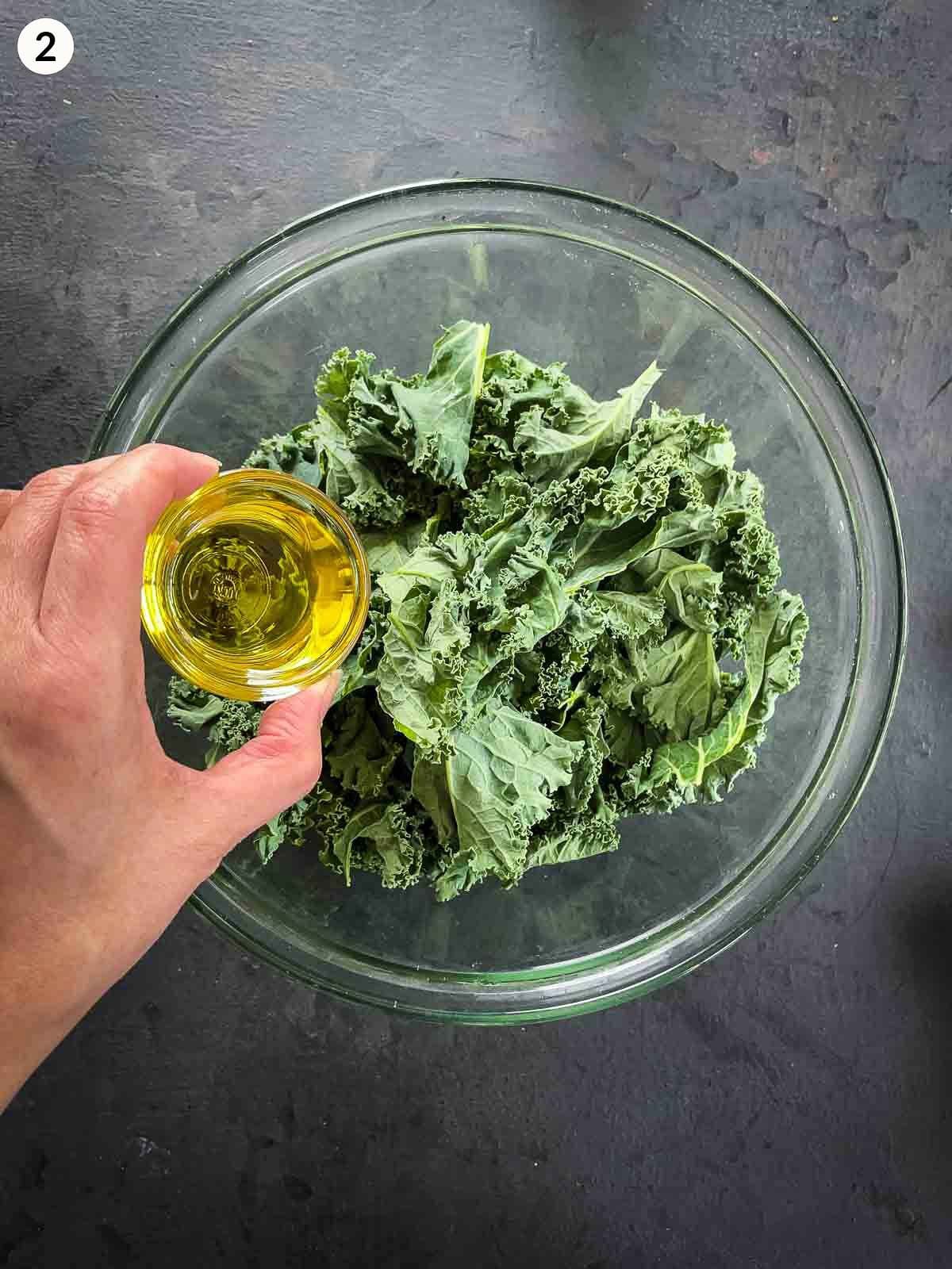 Adding olive oil into into a clear mixing bowl of kale leaves against a dark background