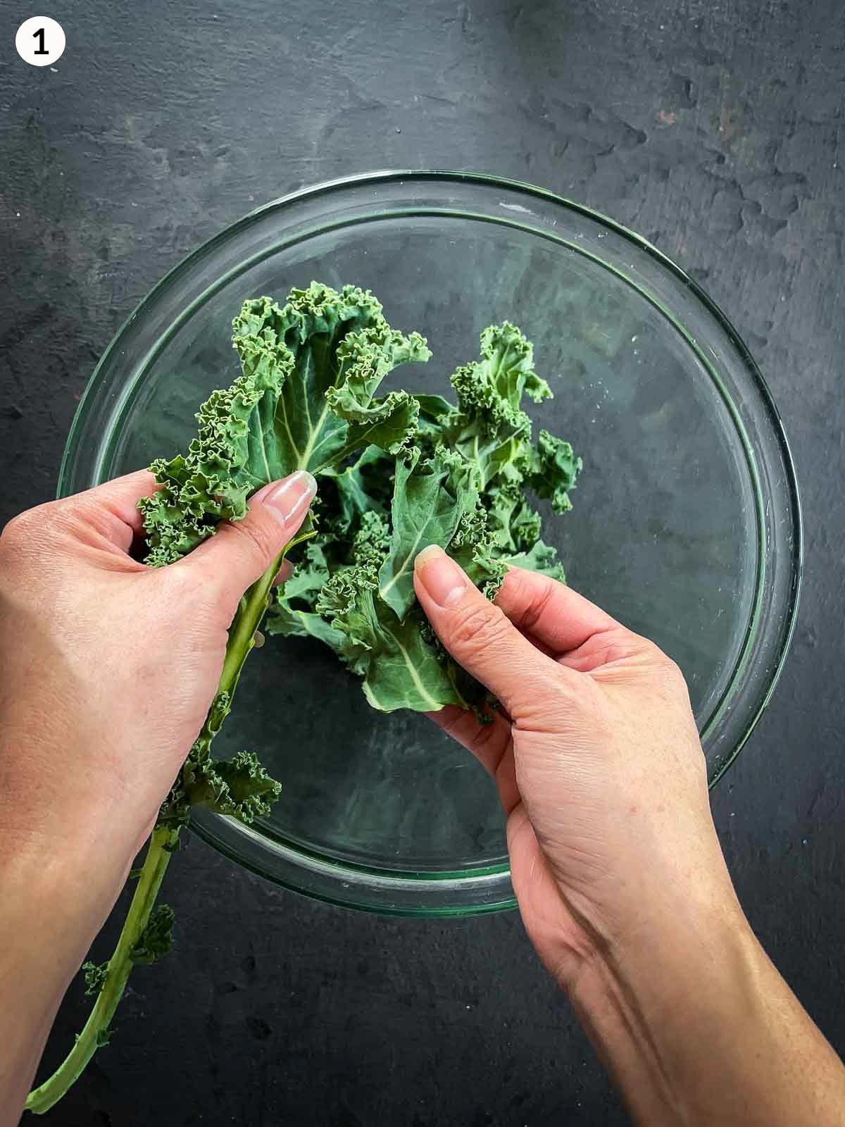 2 hands tearing kale leaves into a clear mixing bowl against a dark background