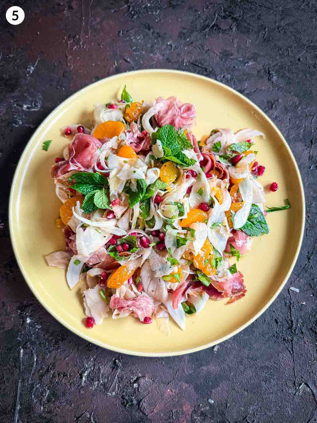 Assembling ingredients for Shaved Fennel Salad with Mandarin, Prosciutto & Pomegranate in a yellow plate
