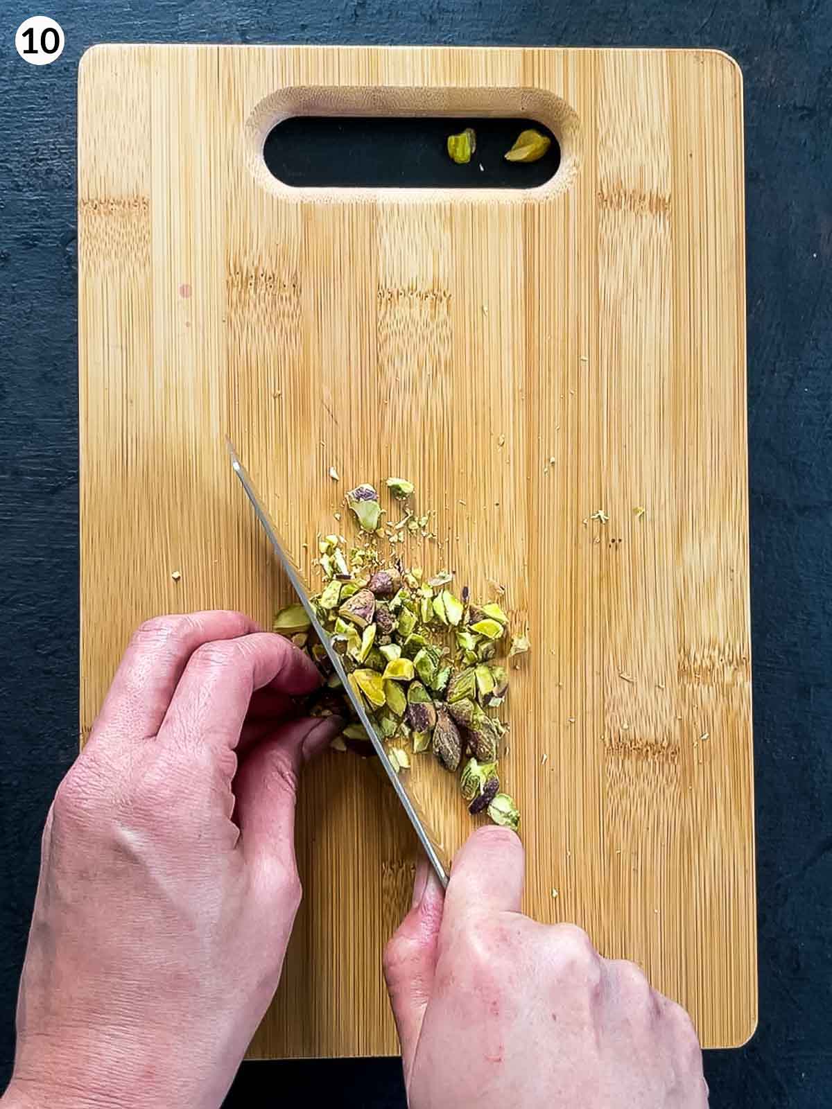Hands cutting pistachios on a wooden chopping board using a chef's knife, against a dark background.