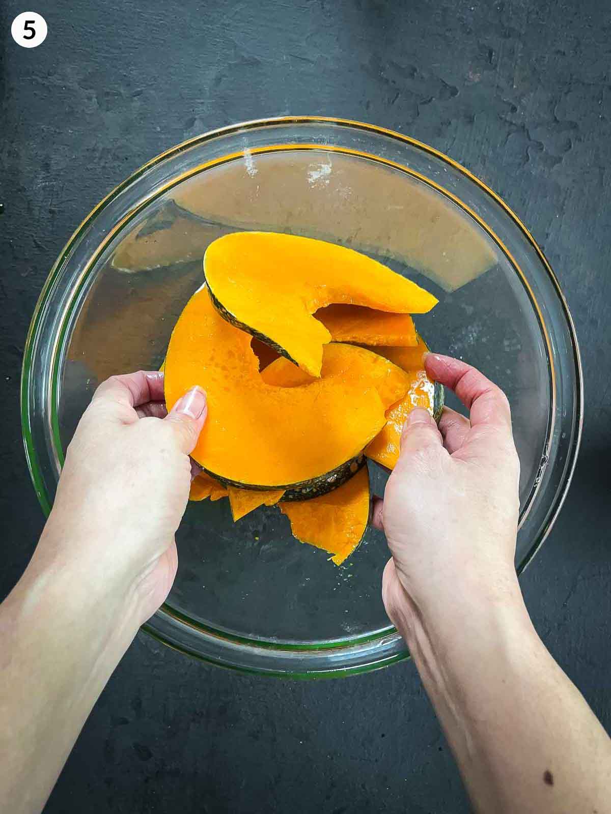 2 hands mixing pumpkin wedges with olive oil in a clear mixing bowl against a dark background