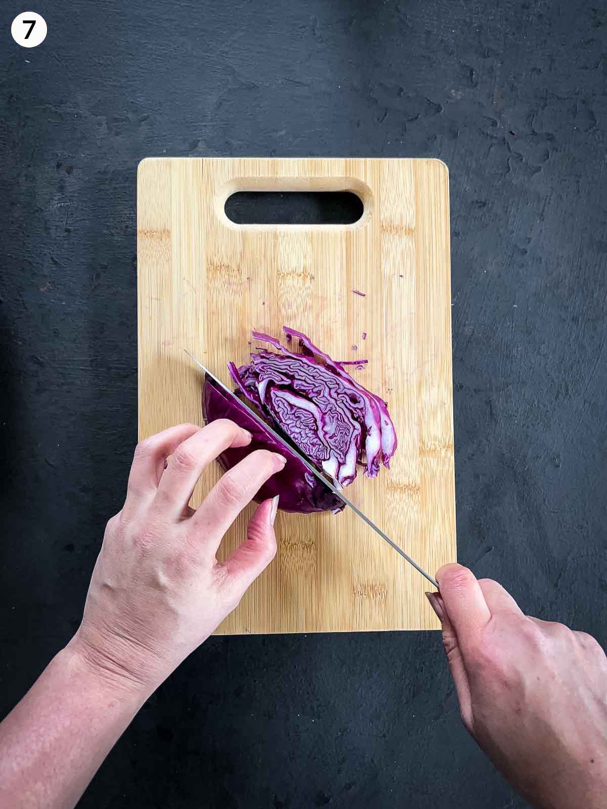 2 hands slicing red cabbage with a knife on a wooden chopping board against a dark background