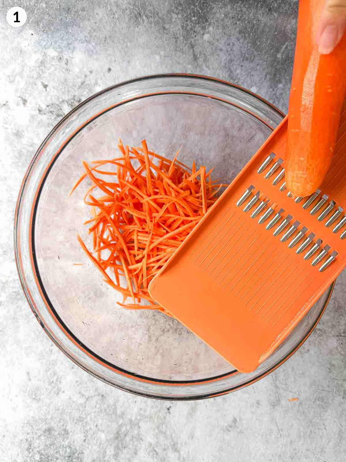 Shredding carrots with an orange coloured mandoline over a clear mixing bowl