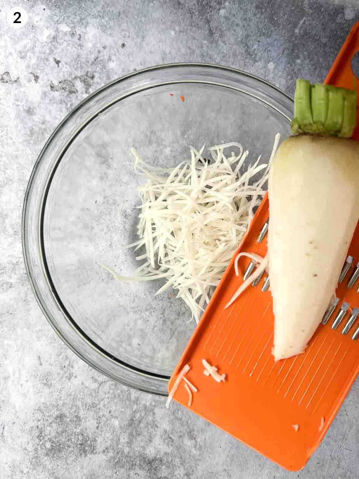 Shredding daikon with an orange coloured mandoline over a clear mixing bowl