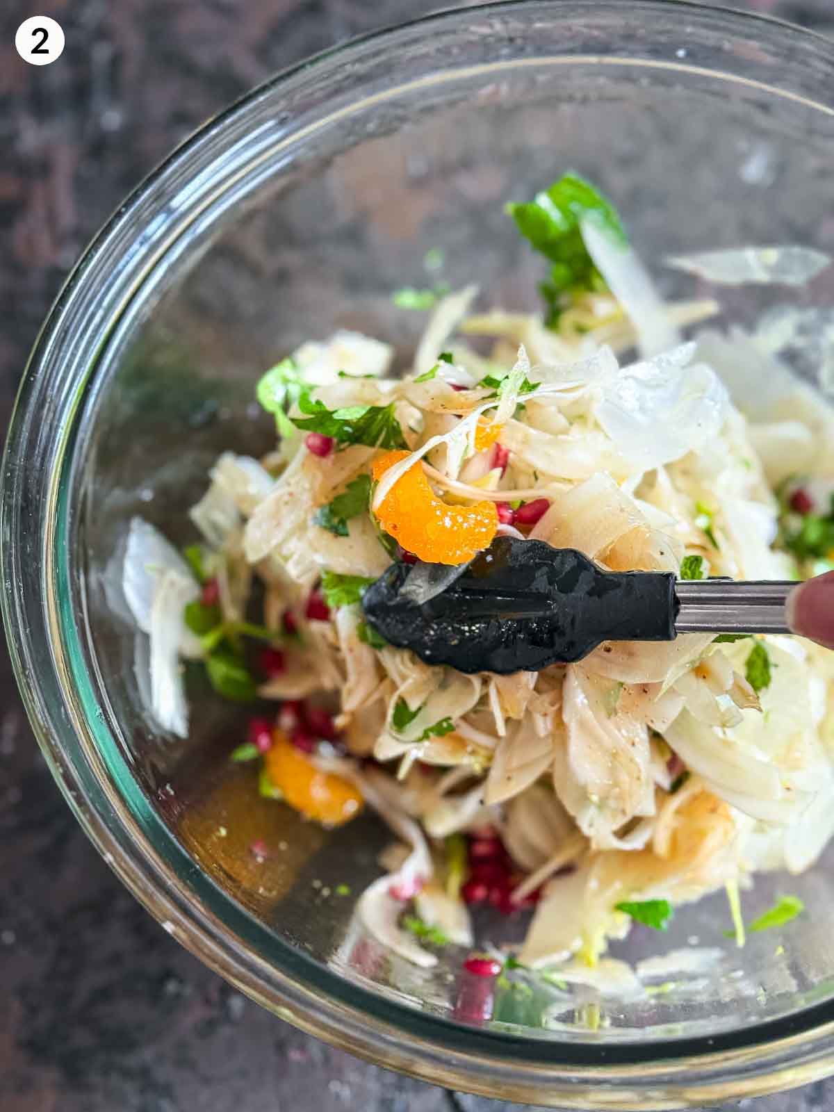 Tossing the ingredients for Shaved Fennel Salad with Mandarin, Prosciutto & Pomegranate with a pair of tongs in a clear bowl