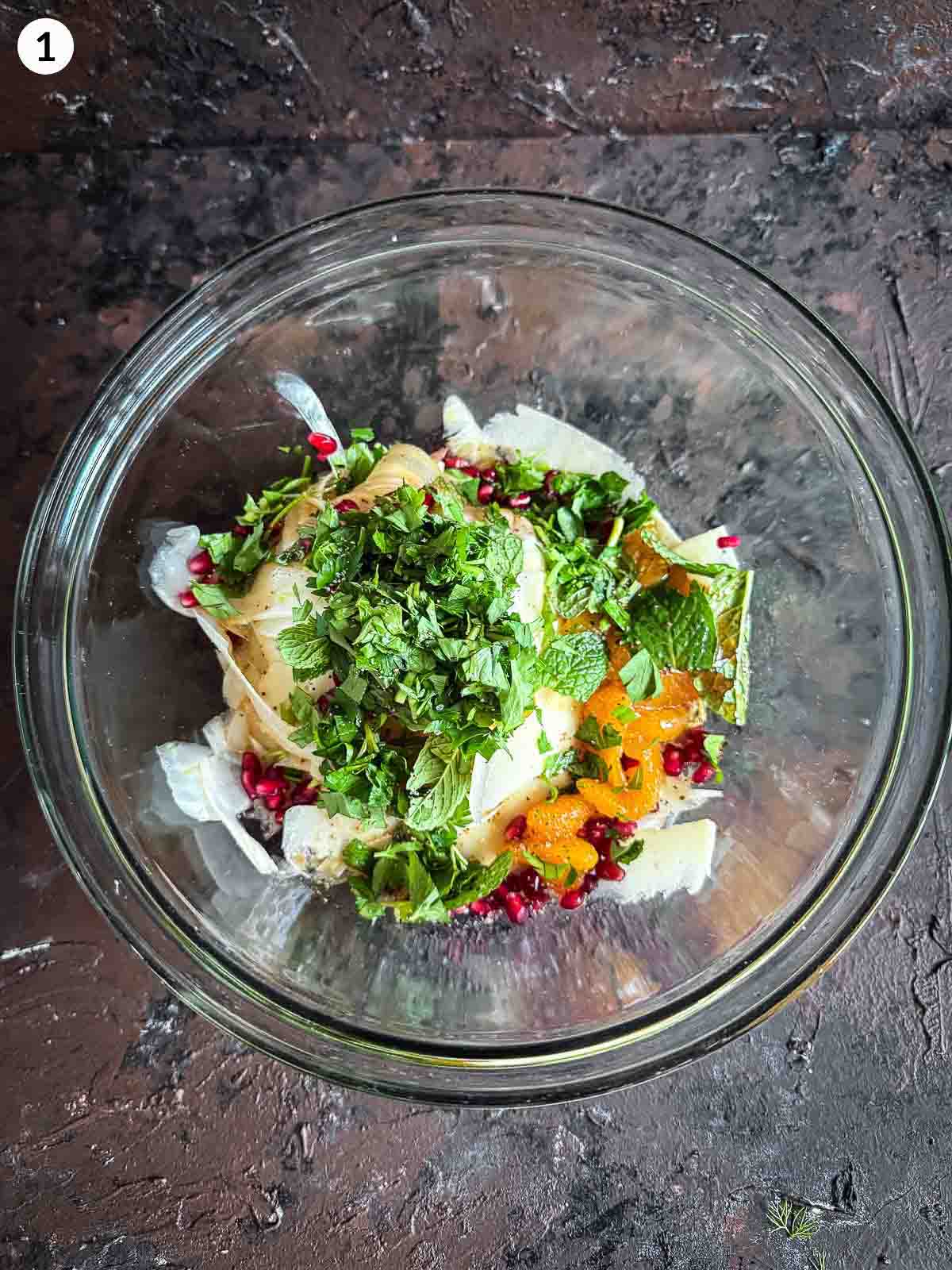 Adding ingredients for Shaved Fennel Salad with Mandarin, Prosciutto & Pomegranate into a clear bowl