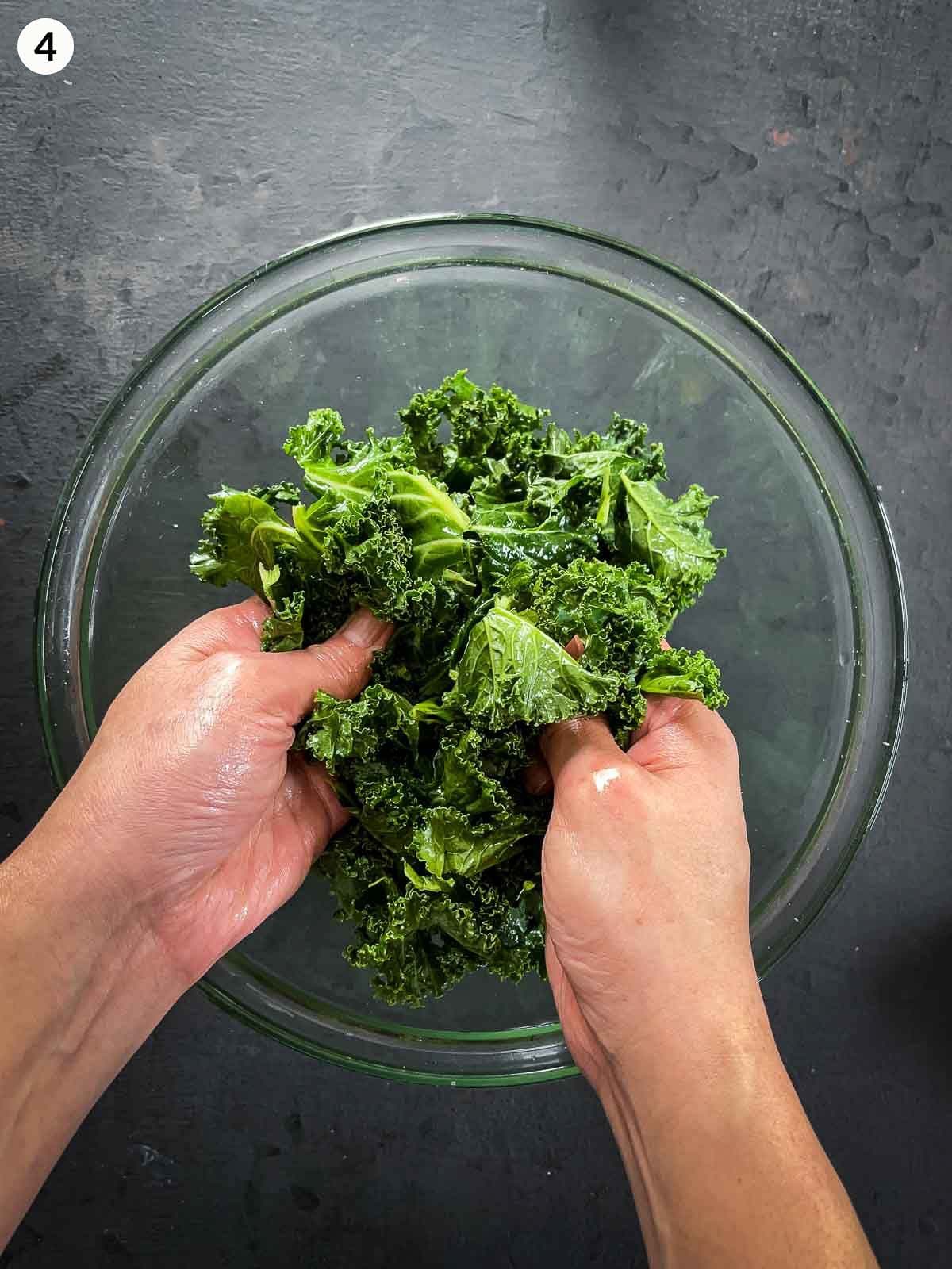 2 hands massaging Adding kale leaves in a clear mixing bowl against a dark background