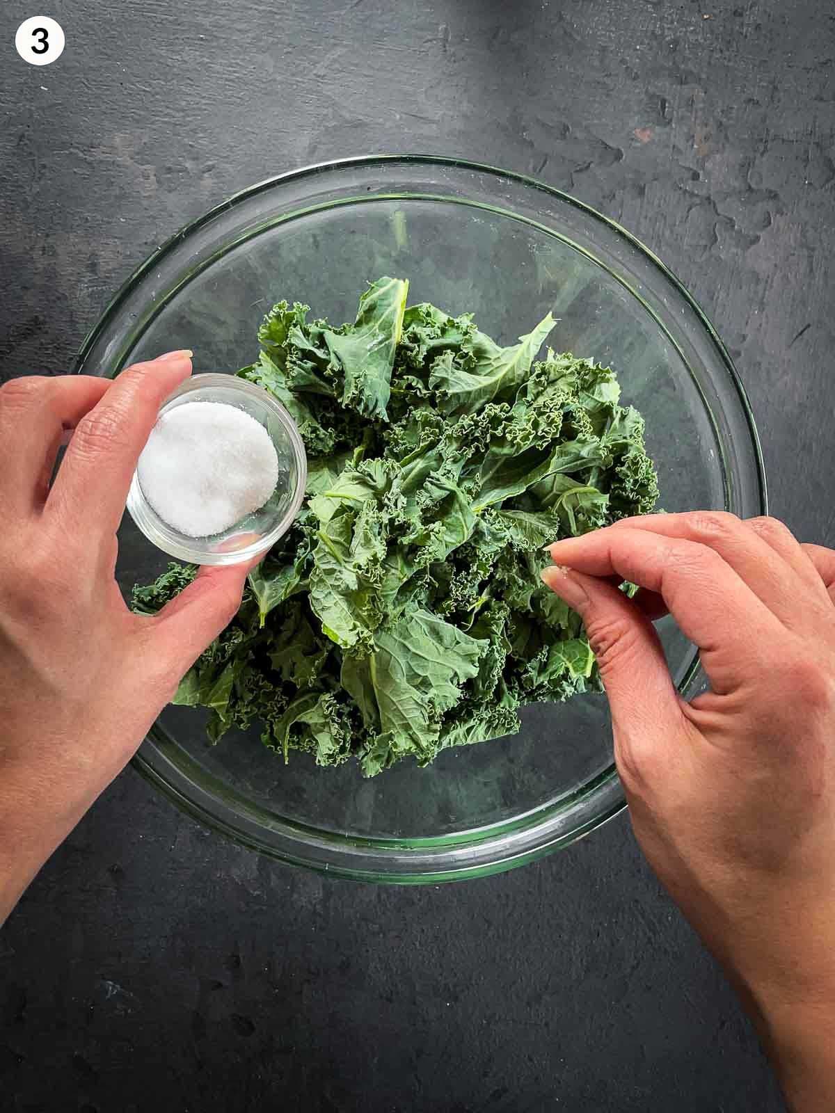 Adding salt into into a clear mixing bowl of kale leaves against a dark background