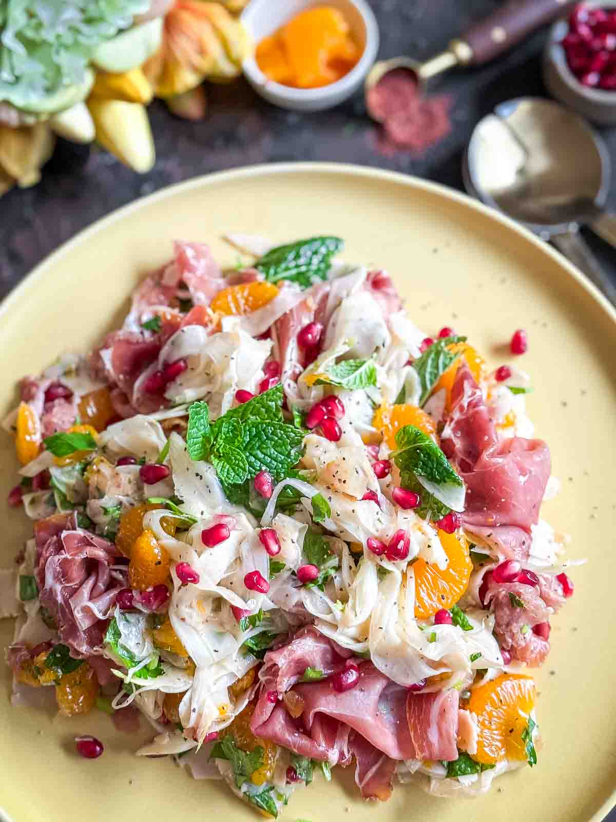 Shaved Fennel Salad with Mandarin, Prosciutto & Pomegranate served on a yellow plate with flowers and spices in the background