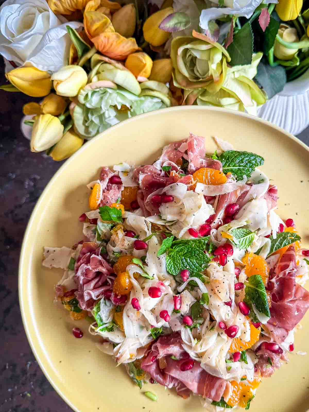 Shaved Fennel Salad with Mandarin, Prosciutto & Pomegranate served on a yellow plate with flowers in the background