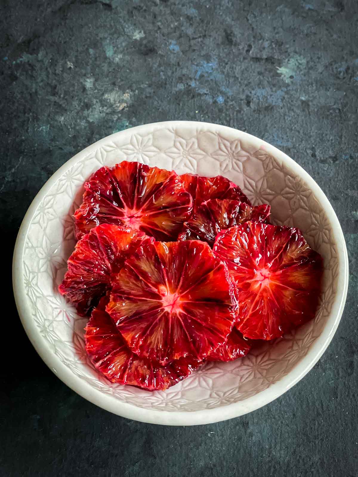 Wheels of sliced blood oranges in a textured bowl against a dark background
