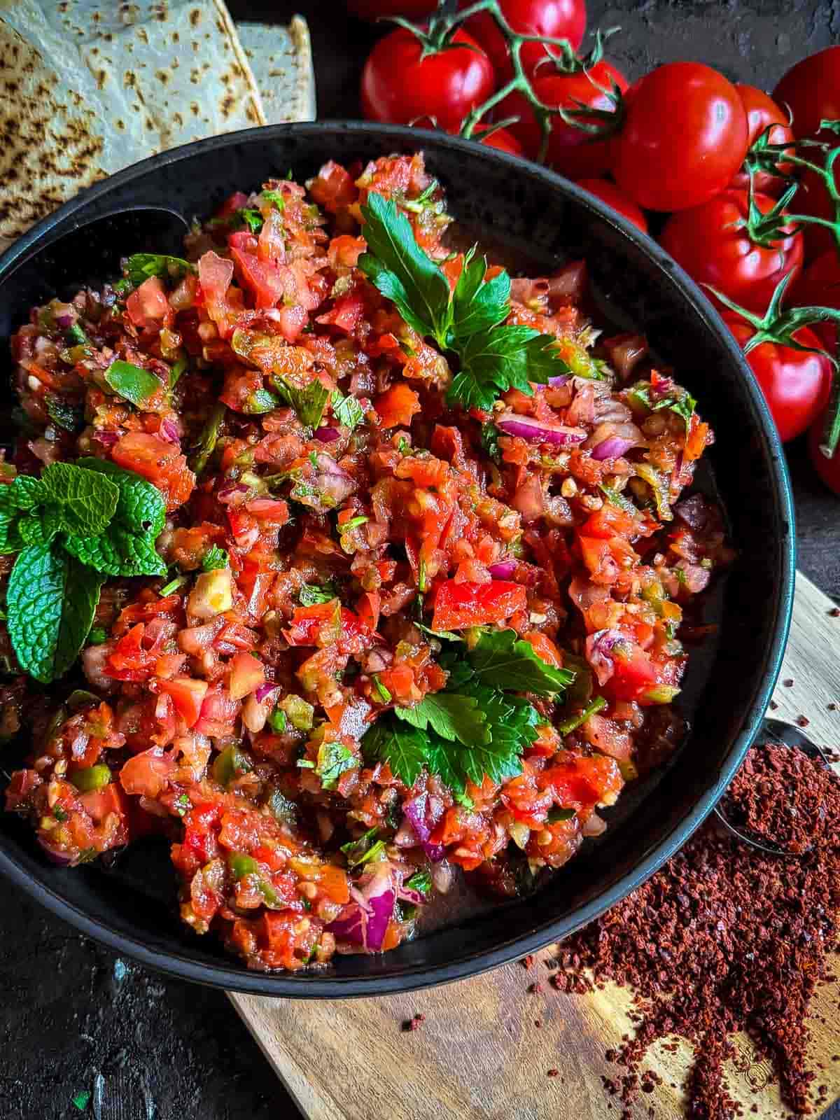 Bowl of Turkish ezme salad in a black plate sitting on a wooden board with Lebanese bread, truss tomatoes and aleppo pepper on the side.