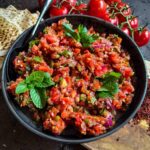 Bowl of Turkish ezme salad in a black plate sitting on a wooden board with Lebanese bread, truss tomatoes and aleppo pepper on the side.