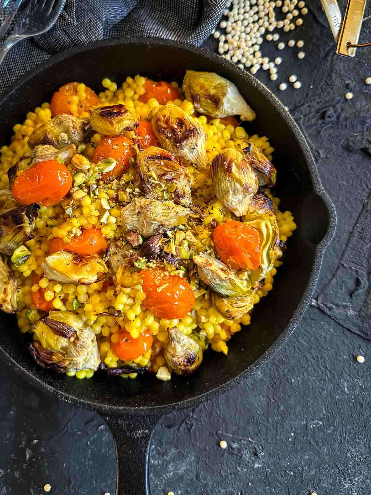 Saffron Israeli Couscous with Roasted Artichoke Hearts in a cast iron pan with blue napkins and uncooked israeli couscous on the side