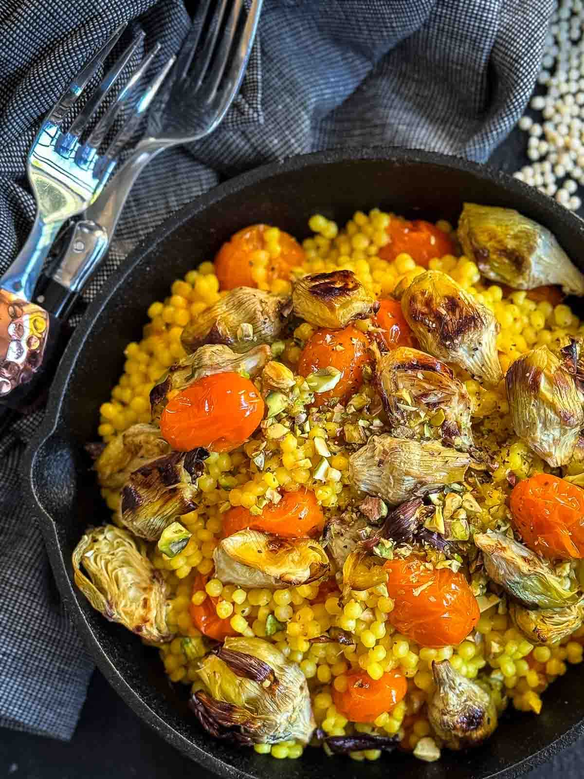 Saffron Israeli Couscous with Roasted Artichoke Hearts in a cast iron pan with blue napkins and cutlery on the side