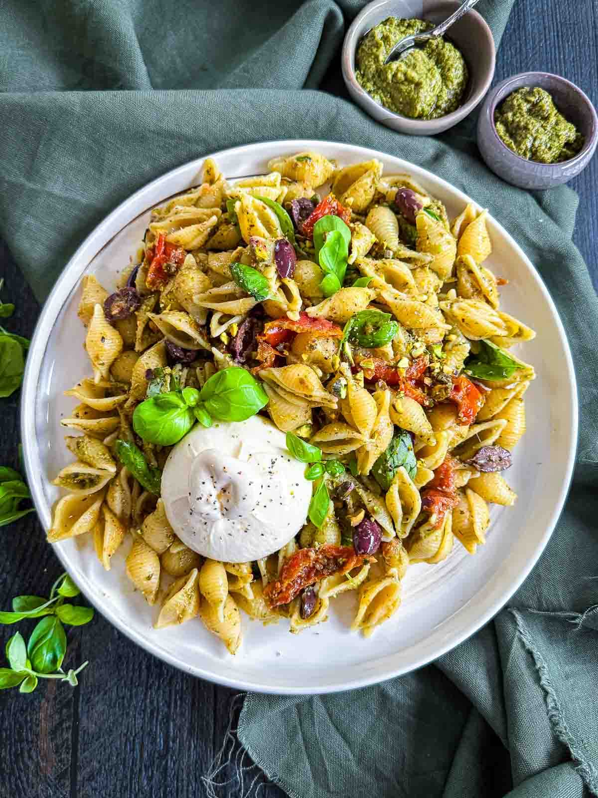 Semi-dried Tomato Pesto Pasta Salad with Burrata on a white plate on green linen cloth with bowls of pesto and basil leaves on the side