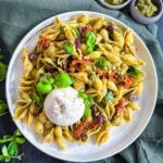 Semi-dried Tomato Pesto Pasta Salad with Burrata on a white plate on green linen cloth with bowls of pesto and basil leaves on the side