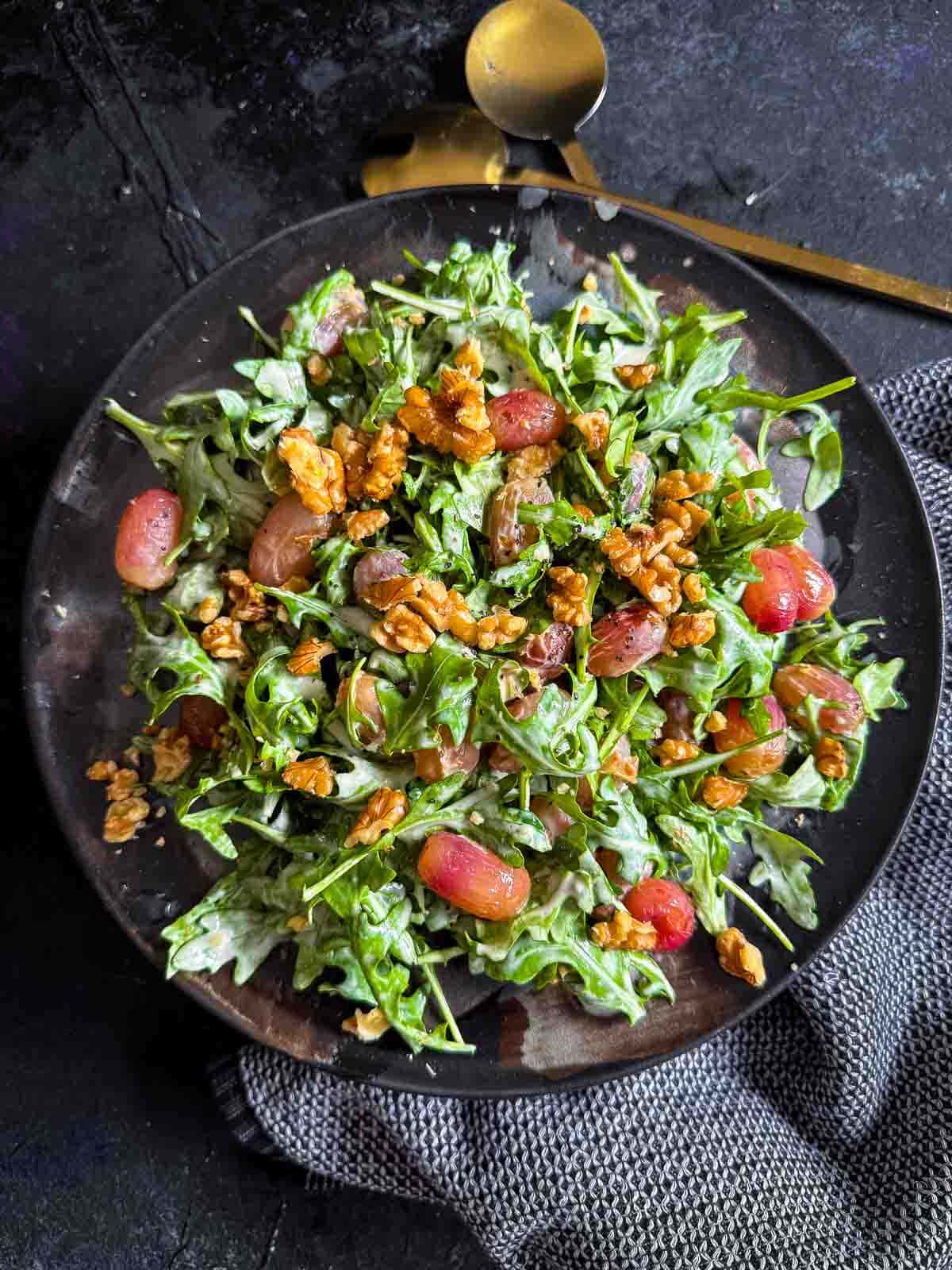 Warm Arugula and Roasted Grape Salad with Blue Cheese served on a dark plate with gold serving cutlery and blue napery on the side