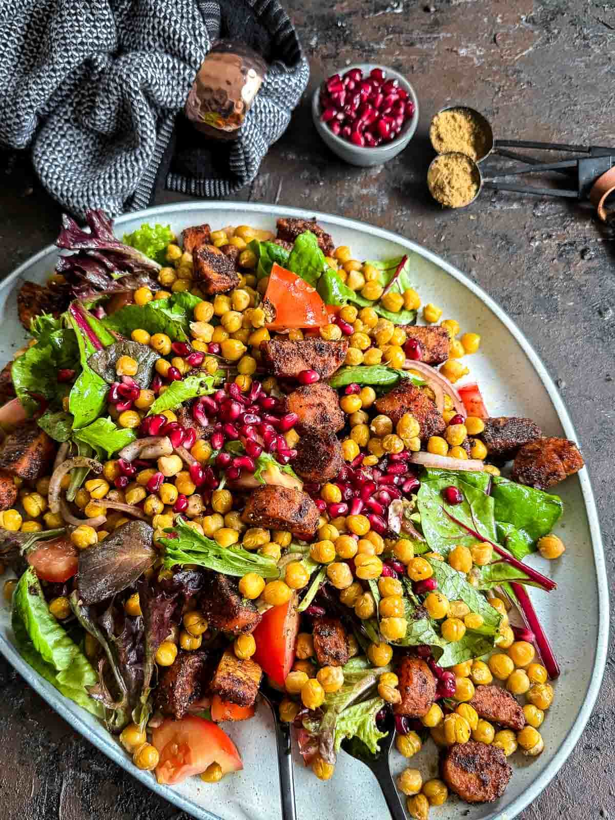 Roasted Chickpea and Carrot Baharat Salad on a grey platter against a dark background with napkin, pomegranate seeds in a bowl and tablespoon of spices