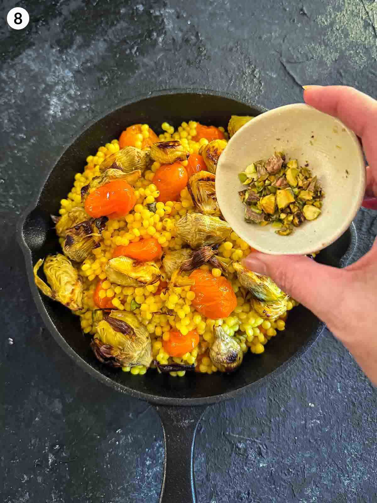 Adding pistachios to Saffron Israeli Couscous with Roasted Artichoke Hearts in a cast iron pan